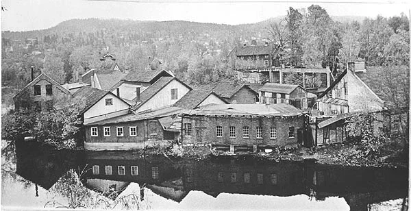 Historisk bilde av sagbruksbygningene tett i tett langs elva, speilet i vannet
